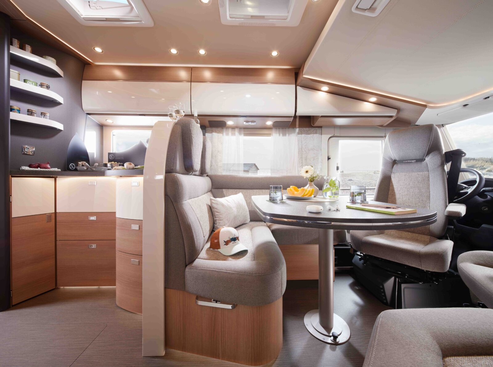 Modernes Wohnmobil-Interieur in Beige und hellem Holz, mit einem Esstisch mit Geschirr, gepolsterten Sitzgelegenheiten, Regalen mit verschiedenen Gegenständen und großen Fenstern, die Tageslicht hereinlassen.