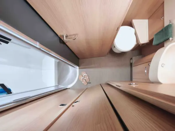 Draufsicht auf ein kleines, modernes Badezimmer in einem vollintegrierten Wohnmobil, mit Holzverkleidung, Dusche mit Glastür, Toilette, kompaktem Waschbecken, Handtuch, Sandalen auf dem Boden und einem praktischen Schrank.