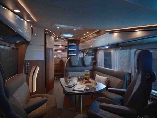 Die luxuriöse reisemobil-Innenausstattung mit moderner Beleuchtung, bequemen Sitzgelegenheiten um einen mit Tee und Snacks gedeckten Tisch, schlichten Schränken und großen Fenstern mit Vorhängen schafft eine gemütliche und einladende Atmosphäre.