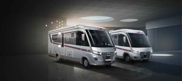 Zwei moderne, silberne, vollintegrierte Wohnmobile mit großen Fenstern und roten Akzentstreifen sind in einem schlichten Raum im Industriestil mit runden Oberlichtern unter dramatischer Beleuchtung geparkt.