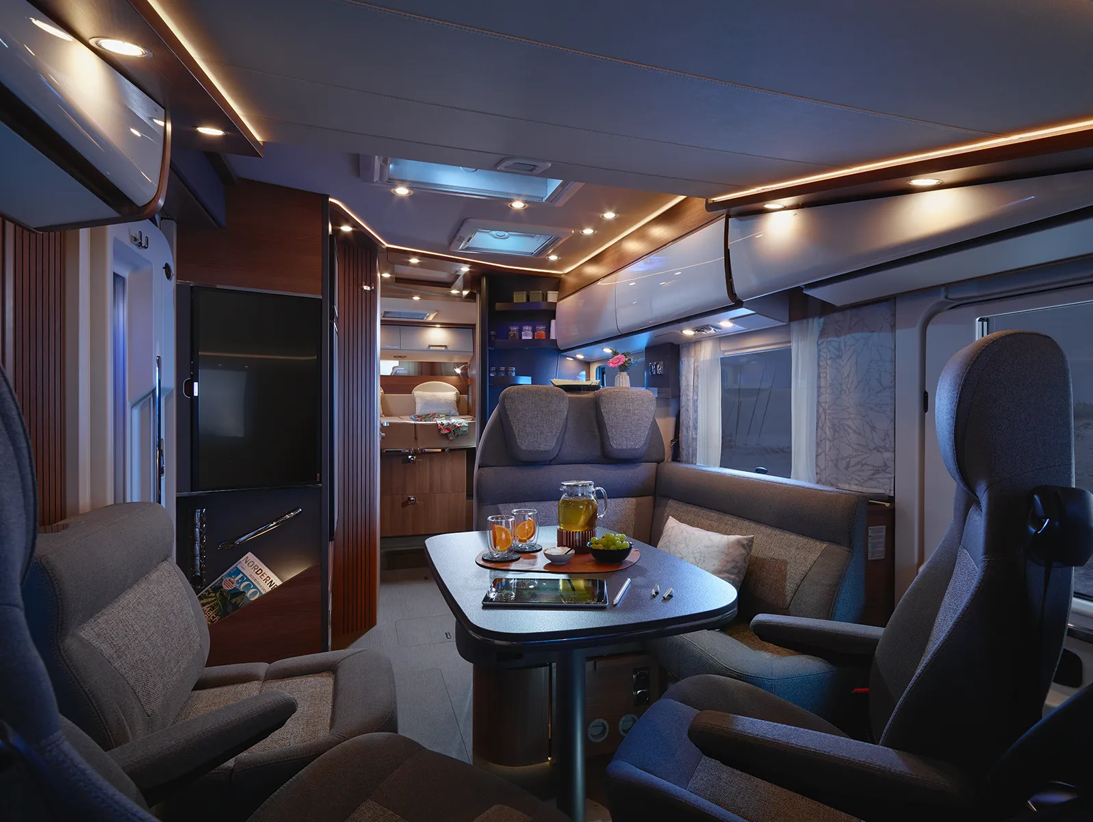 Modernes Carthago Chic C Line Wohnmobil-Interieur mit schlichter, stimmungsvoller Beleuchtung, grauen gepolsterten Sitzen um einen Tisch mit Getränken und Snacks, einer kleinen Küche im Hintergrund und Holzakzenten im gesamten gemütlichen Raum.