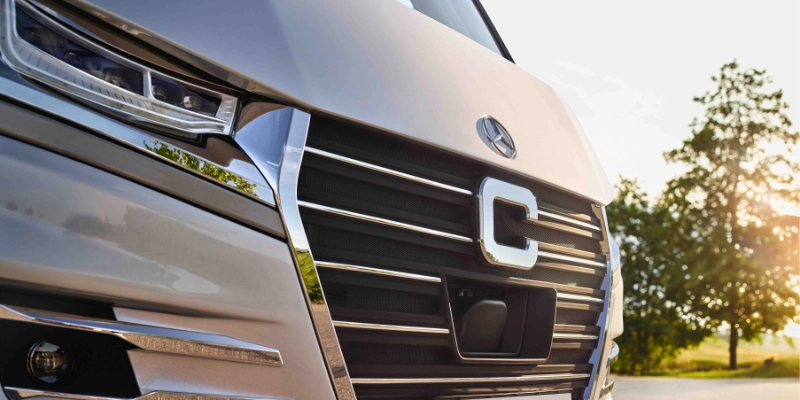 Nahaufnahme des vorderen Kühlergrills und der Scheinwerfer eines modernen silbernen Fahrzeugs mit einem großen "C"-Emblem, aufgenommen im Freien bei Sonnenlicht, wobei Bäume und Himmel im unscharfen Hintergrund zu sehen sind.