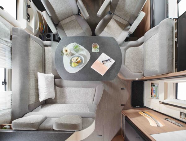 Blick von oben auf einen gemütlichen integrierten wohnmobil-Essbereich mit einem grauen V-förmigen Sofa, einem Tisch mit Snacks, Getränken, einer Blumenvase und einem Notizbuch mit Gläsern. Sanfte Beleuchtung und Regale mit Büchern tragen zum reisemobilen Charme bei.