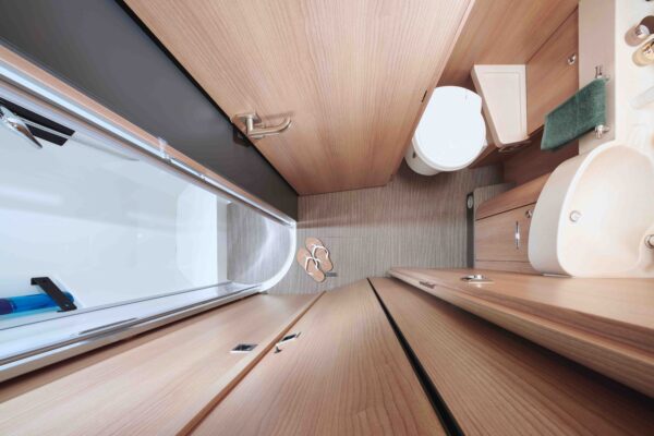Draufsicht auf ein kompaktes wohnmobil-Bad mit Holzverkleidung, Toilette, Waschbecken, Duschbereich mit Glastür, kleinem Schrank, Sandalen auf dem Boden und einem grünen Handtuch an der Wand.