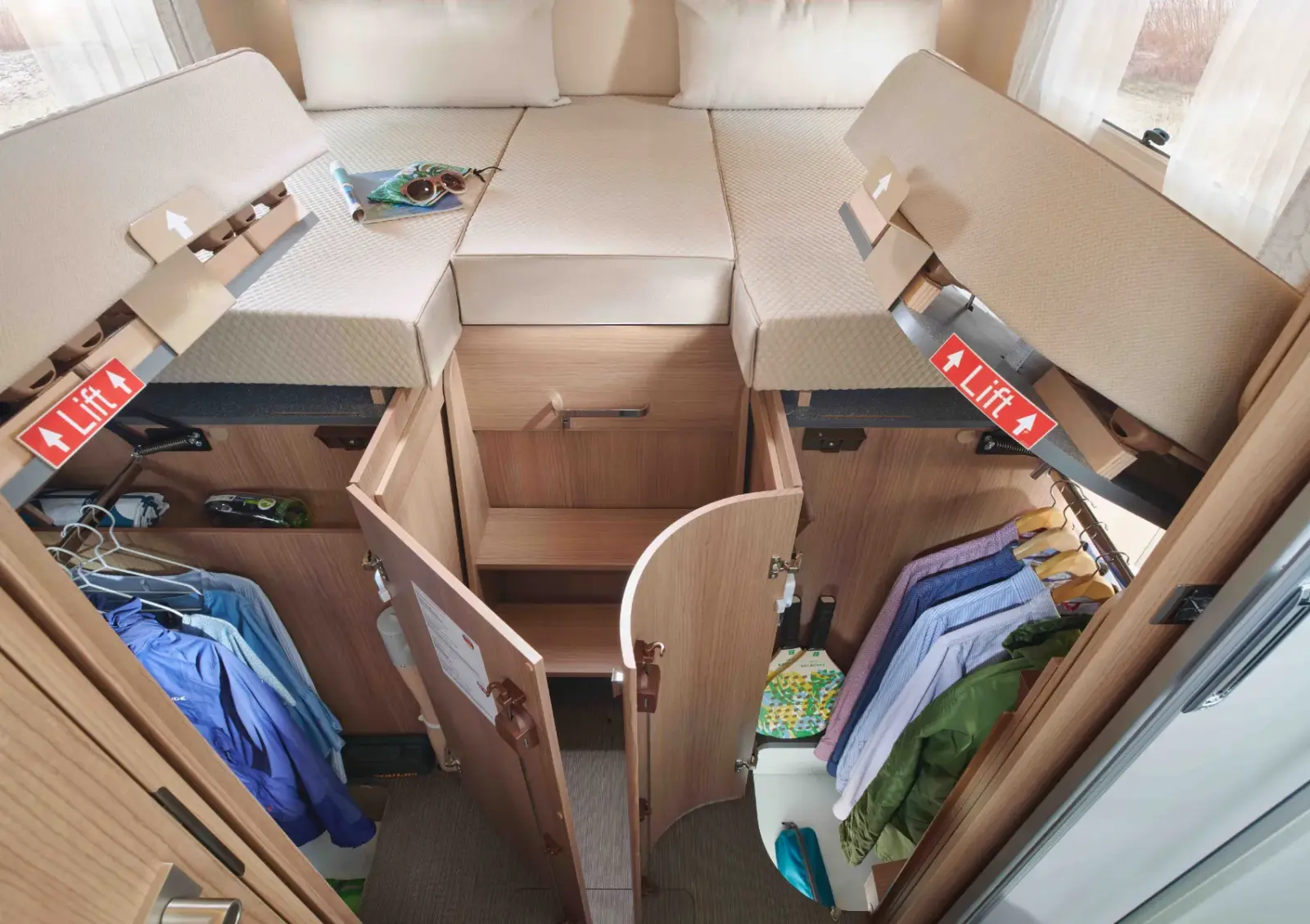 Innenraum eines teilintegrierten Wohnmobil-Schlafzimmers mit zwei erhöhten Einzelbetten; offene Staufächer unter jedem Bett geben den Blick auf Kleidung, Schuhe und Regale frei. Die Schranktüren sind offen und die LIFT-Schilder weisen auf die Stauraumklappen der Betten hin.