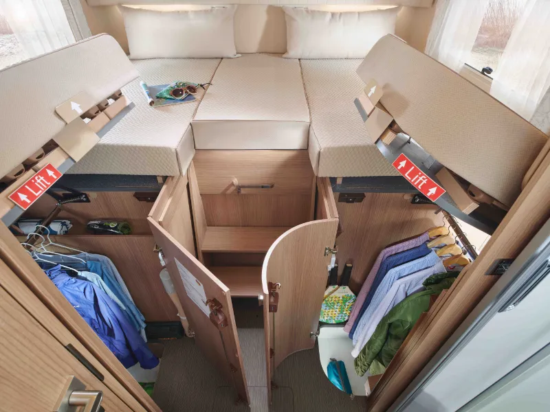 Innenraum eines vollintegrierten Wohnmobils mit zwei Betten, die hochgeklappt werden können, um darunter liegende Staufächer freizugeben. Kleidung hängt ordentlich in Regalen, und persönliche Gegenstände sind in den geräumigen Stauräumen dieses praktischen Reisemobils organisiert.
