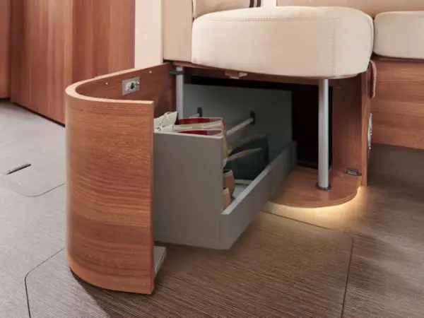 In einem modernen integrierten Wohnmobil wird unter einer beigefarbenen gepolsterten Sitzfläche eine eingebaute Holzschublade herausgezogen, in der sich organisierte Taschen befinden. Die warme Beleuchtung unterstreicht das gemütliche und funktionale Design dieses Wohnraums.
