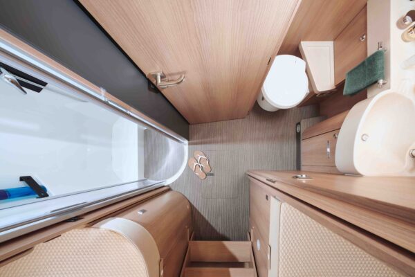 Draufsicht auf ein kompaktes, modernes teilintegriertes Wohnmobil-Bad mit Holzschränken, kleinem Waschbecken, Toilette, Duschbereich, Sandalen auf dem Boden und einem aufgehangenen grünen Handtuch. Der Raum ist aufgeräumt und effizient organisiert.