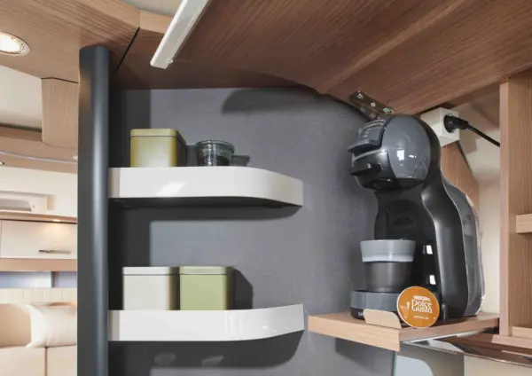 Auf einer modernen Küchentheke in einem integrierten Wohnmobil stehen eine Kapselkaffeemaschine, eine Tasse und eine Kaffeepad auf einem Holzregal. Zwei geschwungene weiße Regale halten Gläser und Tassen vor einer grauen Wand, mit einem gemütlichen Wohnbereich im Hintergrund.