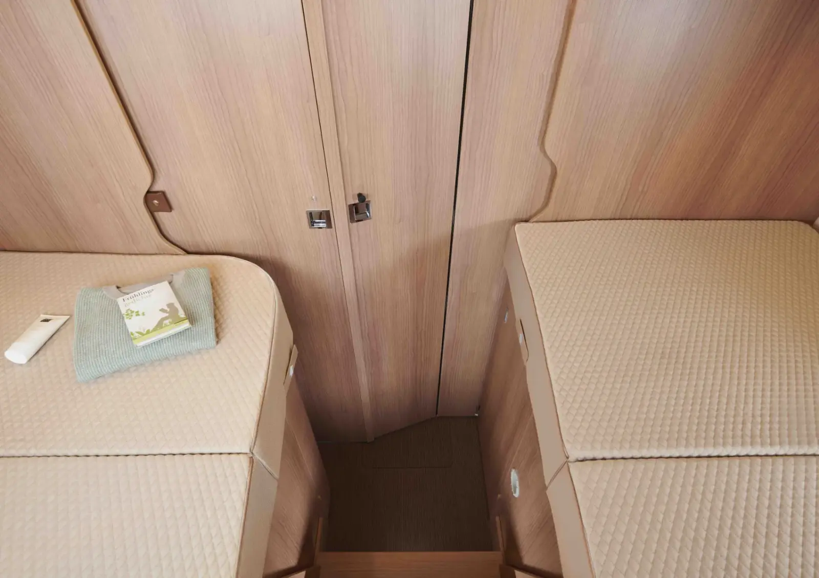 Zwei Einzelbetten mit beigen Steppdecken stehen in diesem teilintegrierten Wohnmobil auf beiden Seiten eines schmalen Holzstegs, der zu einer geschlossenen Holztür führt. Auf einem Bett liegen ein gefaltetes Handtuch und eine Fernbedienung.
