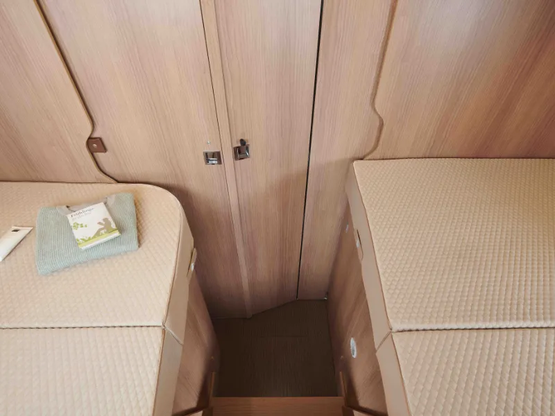 Blick auf zwei Einzelbetten mit beigefarbener Bettwäsche in einem schmalen reisemobil-Zimmer mit Holzwänden und -boden. Auf dem linken Bett liegen ein gefaltetes Handtuch und ein kleines Büchlein, und am anderen Ende befindet sich eine geschlossene Holztür.
