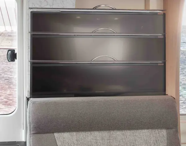 Im Inneren eines modernen Carthago C1 Tourer Plus Teilintegriert stapeln sich vier metallene, an der Wand befestigte Ablagefächer mit geschwungenen Griffen über einem grau gepolsterten Sitz. Licht fällt durch ein Fenster auf der linken Seite ein.