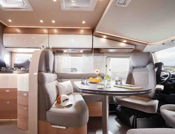 Modernes Carthago C Tourer Wohnmobil-Interieur mit einem Esstisch, gepolsterten Sitzen und großen Fenstern. Auf dem Tisch stehen Gläser, eine kleine Vase mit Blumen und eine Baseballmütze. Helles Holz und neutrale Töne schaffen eine gemütliche, helle Atmosphäre.