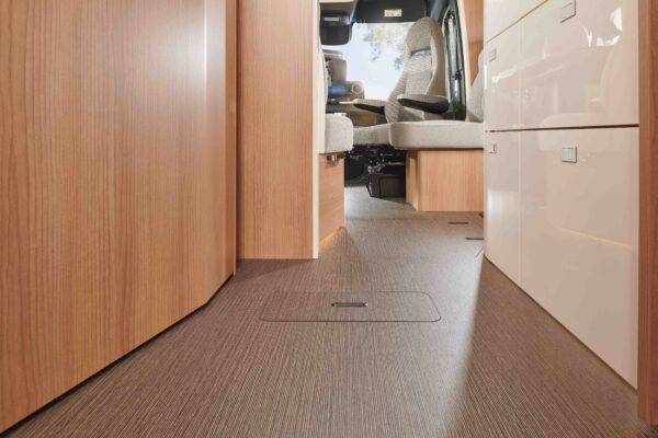 Ansicht eines modernen teilintegrierten Wohnmobils mit holzgetäfelten Schränken auf der linken Seite und beigen Schubladen auf der rechten Seite sowie braunem Bodenbelag, der zu den Fahrer- und Beifahrersitzen im vorderen Bereich führt.