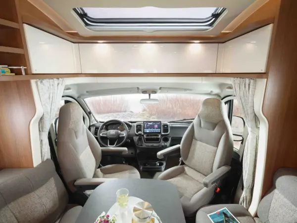 Der Innenraum eines modernen Carthago C1 Tourer Plus teilintegrierten Wohnmobils mit Fahrer- und Beifahrersitz, einem Tisch mit Getränken und Snacks sowie Stauschränken über dem Dach, alles in natürliches Licht getaucht durch ein Oberlicht.