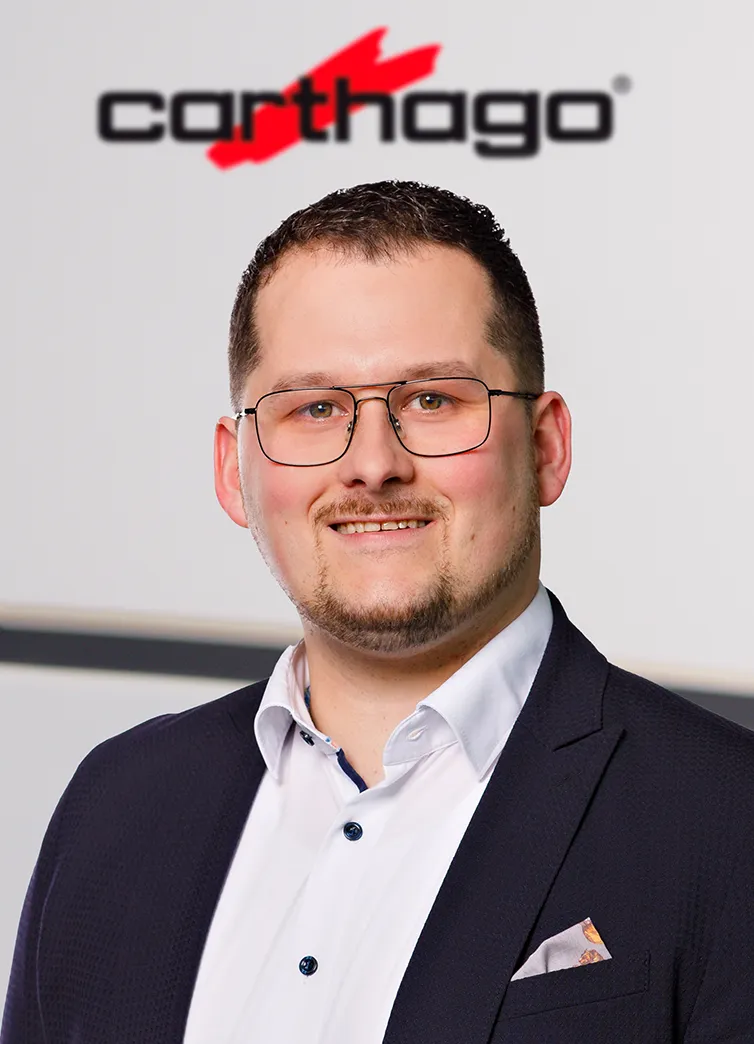 Ein Mann mit kurzen dunklen Haaren, Brille und gestutztem Bart trägt einen marineblauen Anzug und ein weißes Hemd und lächelt vor einer Wand mit dem Carthago Logo im Carthago Werksverkauf.
