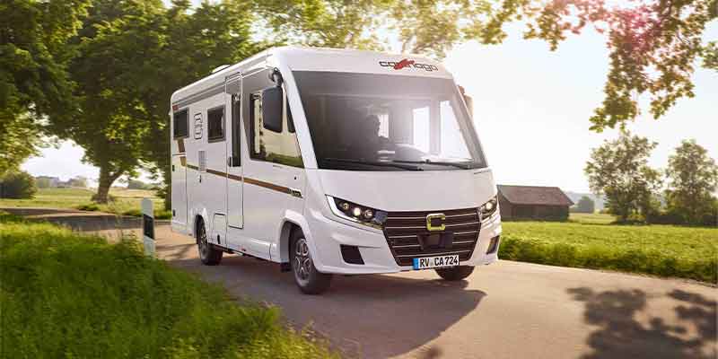Ein weißes Wohnmobil fährt eine Landstraße entlang, umgeben von grünem Gras und Bäumen, durch deren Blätter das Sonnenlicht fällt.