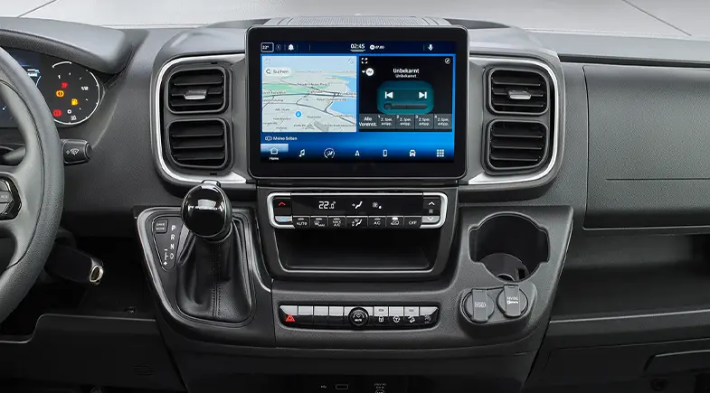 Dashboard of a modern Fiat Ducato motorhome with a large touchscreen display including navigation and media controls, gear lever on the left, aircon buttons at the bottom and cup holder on the right.