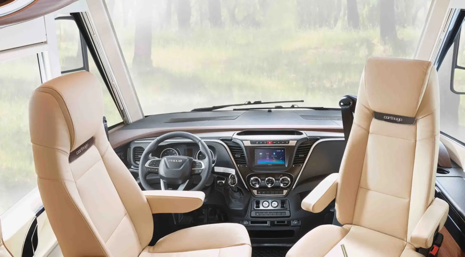 Das Innere eines Wohnmobil-Cockpits mit zwei beigen Kapitänssesseln, einem modernen Armaturenbrett mit Digitalanzeige, Lenkrad und großen Fenstern, die einen verschwommenen waldähnlichen Hintergrund zeigen.