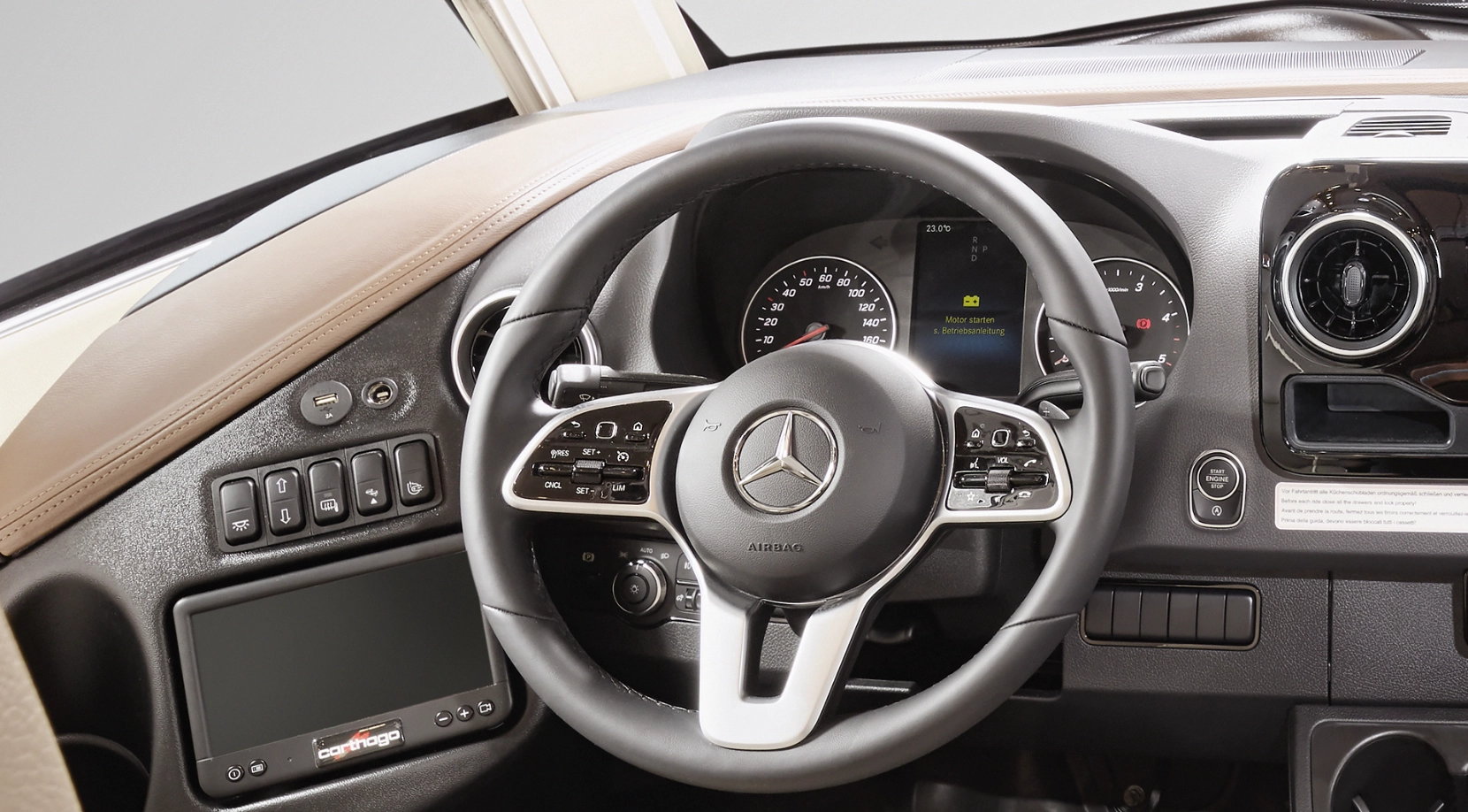 Nahaufnahme eines Mercedes-Benz-Fahrzeuglenkrads mit montierten Bedienelementen, Armaturenbrettanzeigen, Infotainment-Display, Tasten auf der linken Seite und kreisförmigen Lüftungsschlitzen, die ein modernes, luxuriöses Fahrzeuginterieur zeigen.