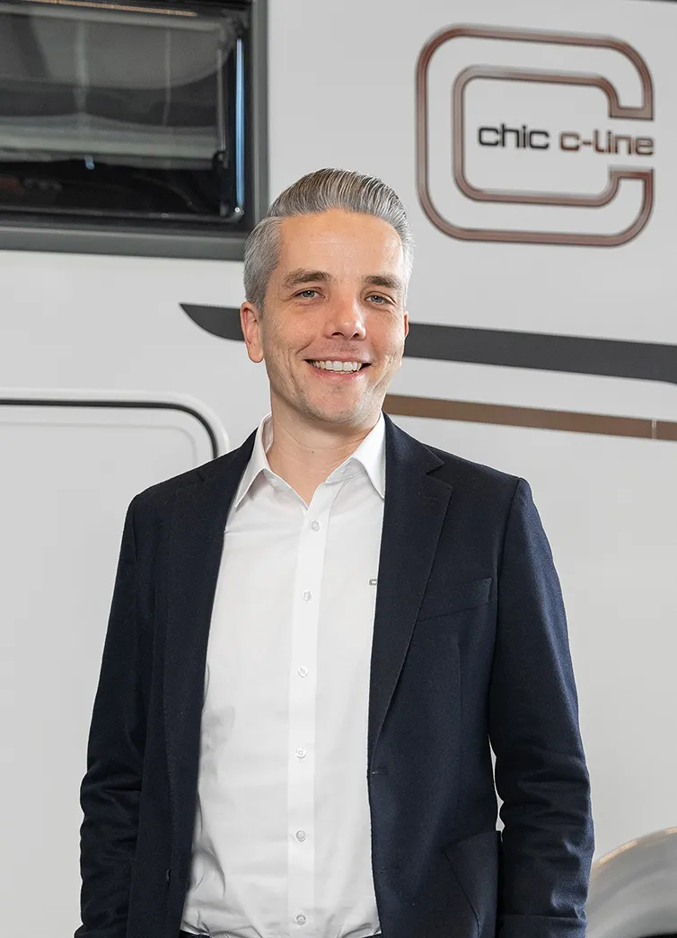 Ein lächelnder Mann in weißem Hemd und dunklem Blazer steht vor einem Fahrzeug mit dem schicken c-line-Logo, das auf die exklusive Carthago Werksverkaufsveranstaltung hinweist.