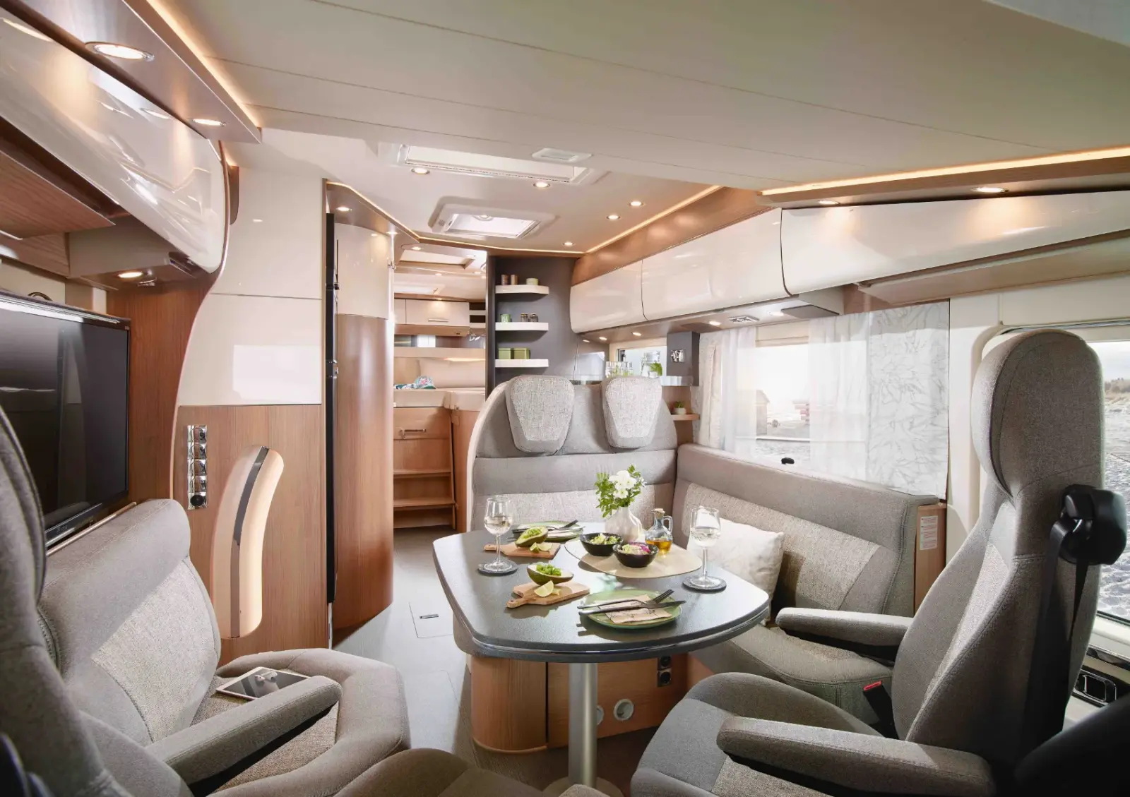Modernes integriertes Wohnmobil-Interieur mit gepolsterten Sitzen um einen mit Tellern, Schalen und Gläsern gedeckten Esstisch. Helle Holzschränke, Regale und eine sanfte Beleuchtung schaffen eine gemütliche Atmosphäre. Eine kleine Vase mit Blumen steht auf dem Tisch.
