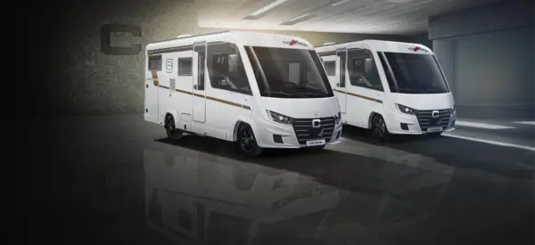 Zwei weiße, moderne Wohnmobile vom Typ Carthago C2-Tourer mit schwarzen Akzenten sind in einem Innenraum auf einem spiegelnden Boden bei gedämpfter Beleuchtung geparkt, mit Blickrichtung nach links, mit Betonwänden und einem teilweise geöffneten Garagentor im Hintergrund.