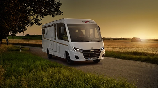 Ein weißes Carthago-Wohnmobil fährt bei Sonnenuntergang über eine Landstraße in Slowenien, umgeben von Feldern und Bäumen, mit einem warmen orangefarbenen Himmel im Hintergrund.
