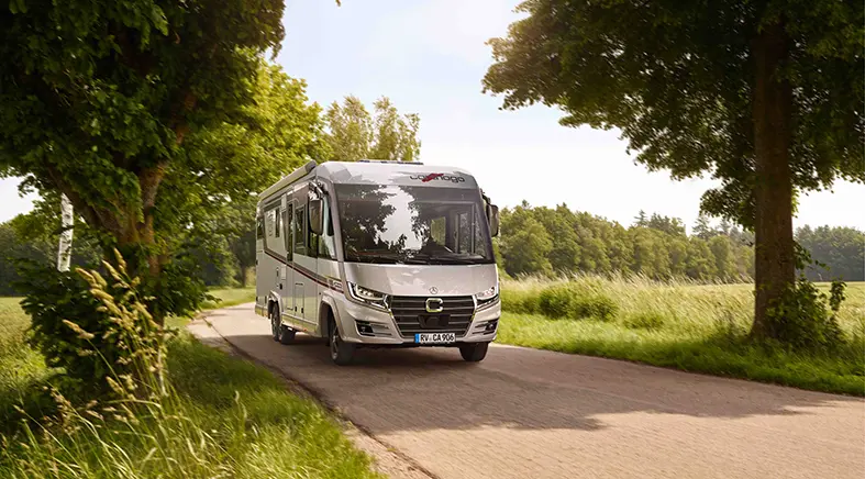 Ein silbernes Carthago-Wohnmobil fährt auf einer schmalen Landstraße in Slowenien, umgeben von grünen Bäumen und grasbewachsenen Feldern unter einem teilweise bewölkten Himmel.