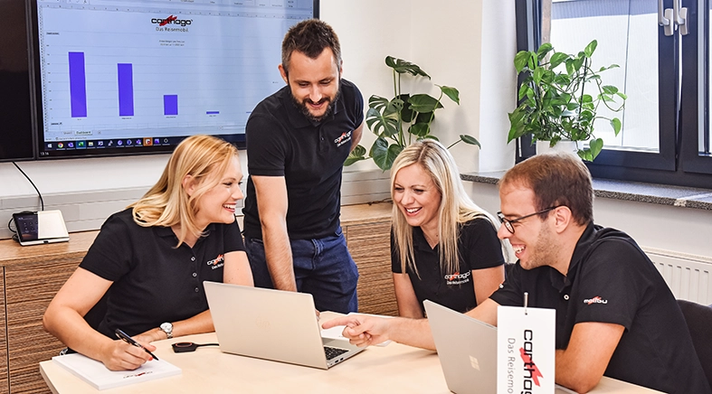 Vier Kollegen in passenden schwarzen Hemden sitzen und stehen um einen Tisch herum, lächeln und arbeiten an Laptops. Auf einem großen Bildschirm im Hintergrund ist ein Balkendiagramm zu sehen, das Daten aus dem Projekt Carthago Slowenien zeigt.