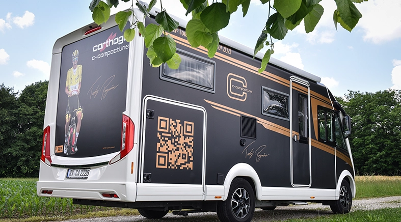 Ein schwarz-weißes Carthago-Wohnmobil mit goldenen Akzenten ist auf einem Kiesweg in der Nähe eines Feldes in Slowenien geparkt. Das Fahrzeug ist mit einem großen QR-Code, einer Radfahrergrafik und Unterschriften auf der Seite und der Rückseite versehen. Darüber hängen grüne Baumblätter.