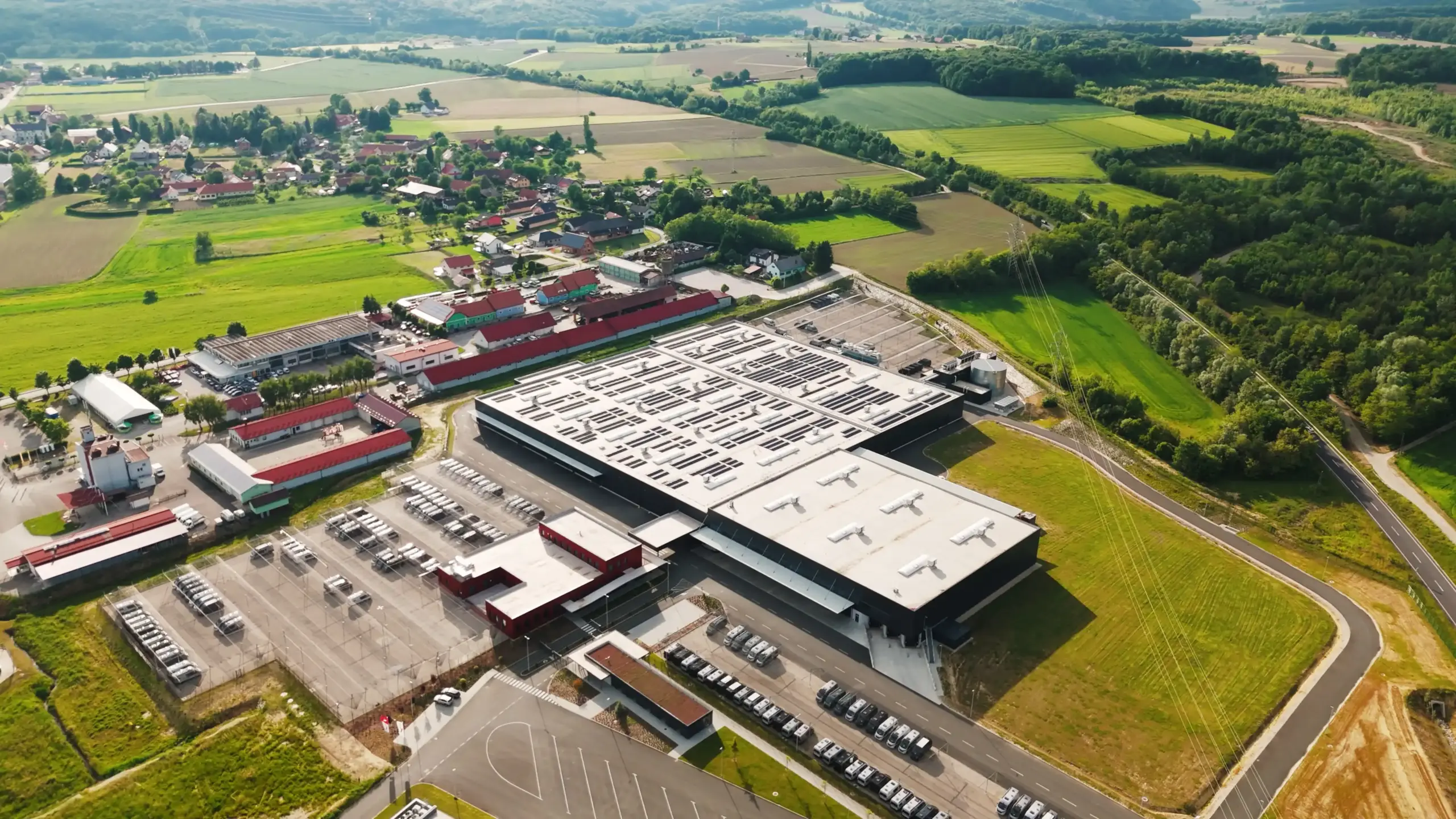 Luftaufnahme eines großen Industriekomplexes von Carthago Slowenien mit einem flachen, weißen Dach und mehreren Nebengebäuden, umgeben von grünen Feldern und einem kleinen ländlichen Dorf im Hintergrund. Zahlreiche Autos sind vor dem Gebäude geparkt.