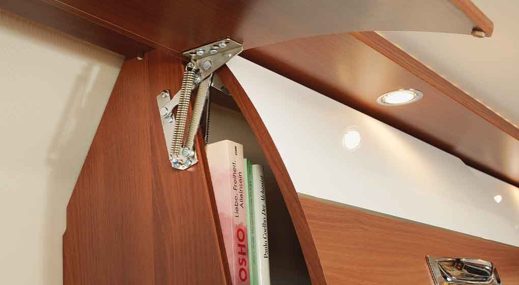 Nahaufnahme einer hölzernen Schranktür mit einem Metallscharnier und einem Federmechanismus, die teilweise geöffnet ist und mehrere Bücher im Inneren offenbart. In der oberen Platte über dem Schrank sind Einbaulampen installiert.