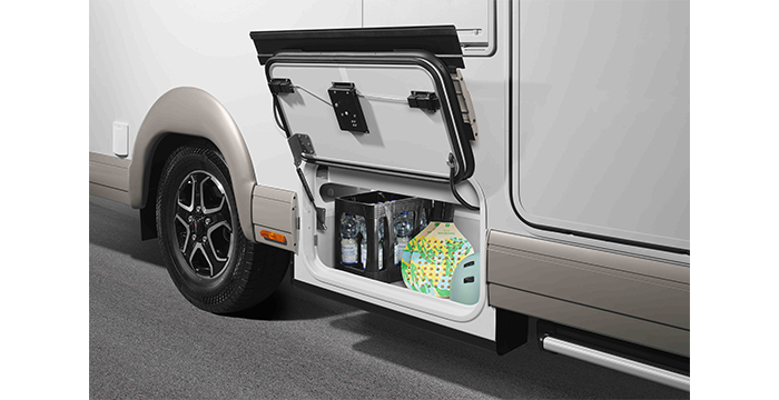 Eine Nahaufnahme des äußeren Seitenfachs eines Wohnmobils, das geöffnet wurde, um Gegenstände wie Wasserflaschen, eine Packung Papierhandtücher und einen grün-weißen Behälter zu zeigen. Das Wohnmobil ist auf einer gepflasterten Fläche geparkt.