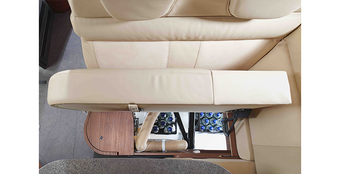 Draufsicht auf ein beigefarbenes Ledersofa in einem Wohnmobil, mit einem Staufach unter dem Sitz, in dem mehrere Wasserflaschen aufbewahrt werden. Holzverkleidung und ein Teil einer Arbeitsplatte sind ebenfalls zu sehen.