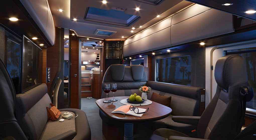 Luxuriöses Wohnmobil-Interieur mit moderner Beleuchtung, Ledersitzen, einem runden Tisch mit Weingläsern, Obst und einem Buch. Der Raum verfügt über große Fenster, Stauraum über Kopf und ein komfortables, einladendes Ambiente.