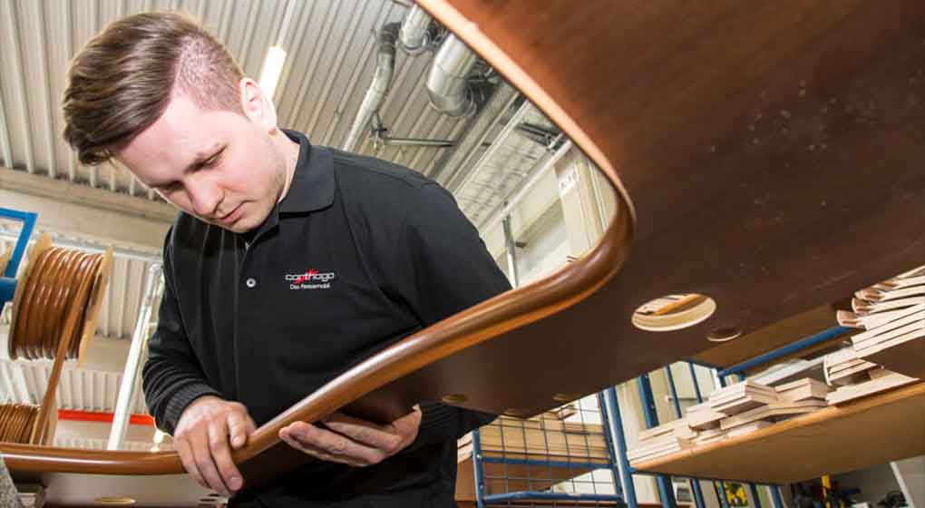 Eine Person in einem schwarzen Hemd prüft eine gebogene Holzplatte in einer Werkstatt, die mit Holzteilen und Industriebeleuchtung gefüllt ist.