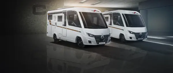 Due moderni camper bianchi, tra cui un Carthago C2-Tourer dal design slanciato, sono parcheggiati uno accanto all’altro in uno spazio interno e sono illuminati da luci intense per mettere in risalto le loro caratteristiche esterne e il marchio.