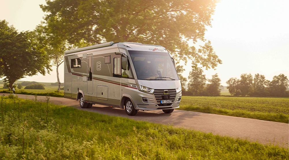 Ein Iveco Daily Wohnmobil fährt eine schmale Landstraße hinunter, umgeben von grünen Feldern und Bäumen, und das Sonnenlicht, das durch den Himmel fällt, schafft eine warme und heitere Atmosphäre.