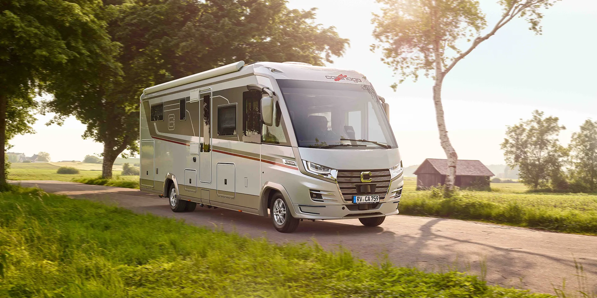 Ein modernes weißes Wohnmobil vom Typ Carthago Chic S Plus fährt an einem sonnigen Tag eine Landstraße entlang, umgeben von grünem Gras, Bäumen und einer kleinen Holzhütte im Hintergrund.