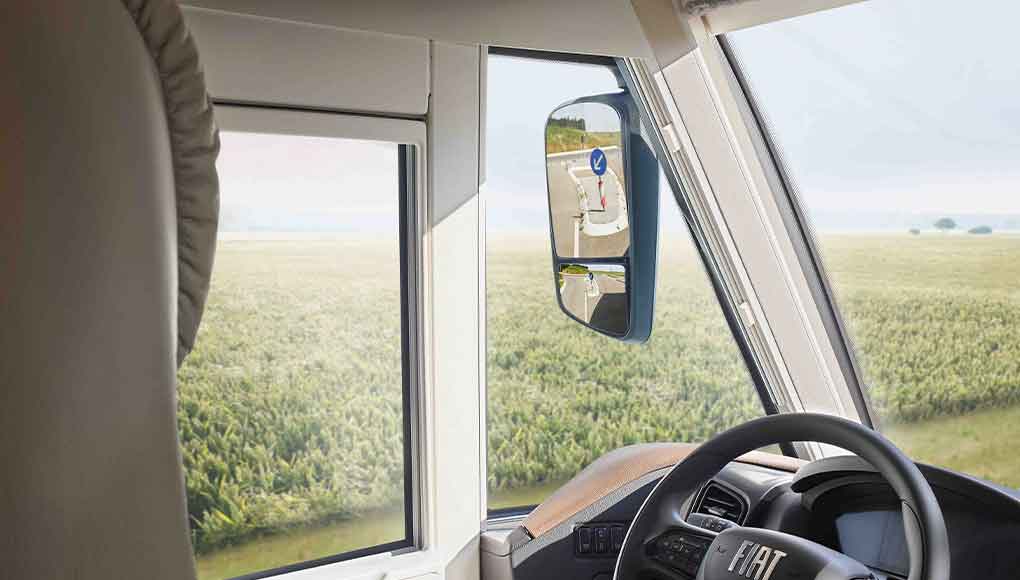 Blick aus dem Inneren eines Fiat-Fahrzeugs mit Lenkrad, Seitenfenster und Seitenspiegel, in denen sich ein Verkehrsschild spiegelt. Draußen ist eine grüne Wiese und ein offener Himmel zu sehen.