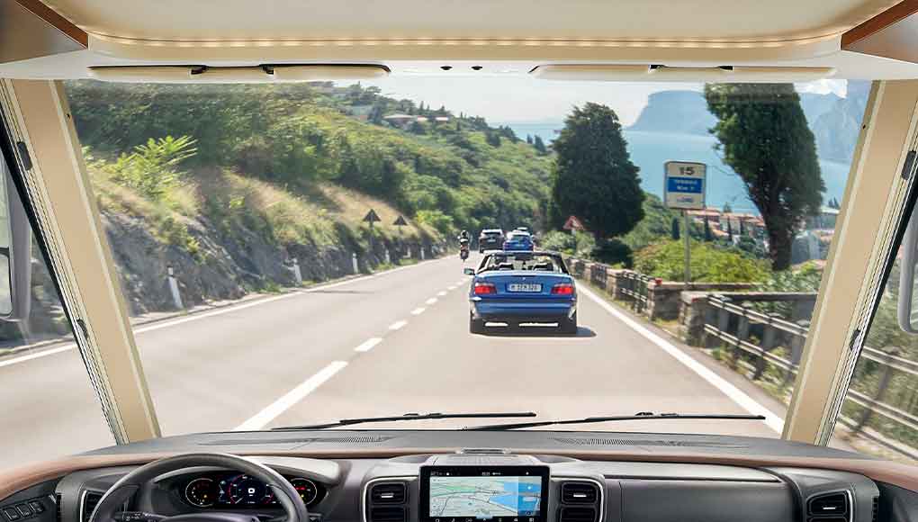Blick aus dem Inneren eines Fahrzeugs durch die Windschutzscheibe auf eine malerische, kurvenreiche, von Bäumen und Geländern gesäumte Straße, auf der mehrere Autos fahren und in der Ferne ein Gewässer zu sehen ist.