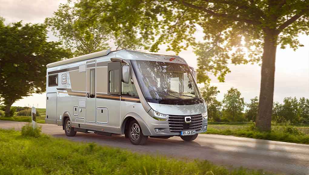 Ein silbernes Wohnmobil fährt eine Landstraße hinunter, die von grünem Gras und Bäumen gesäumt ist, durch deren Äste das Sonnenlicht fällt.