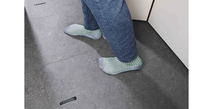 Person mit hellgrün-grau gestreiften Socken und blauer Hose, die auf einem dunkelgrauen Kachelboden neben einem hellen Schrank steht.