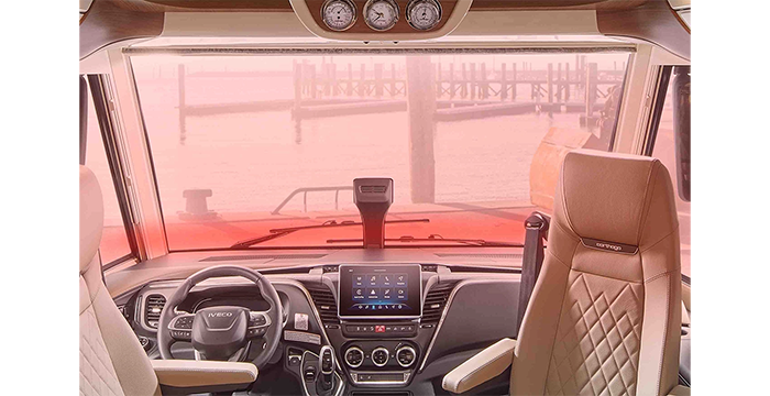 Innenansicht eines modernen Wohnmobil-Cockpits mit zwei hellbraunen Ledersitzen, einem Lenkrad, einem Armaturenbrett mit digitalen Bedienelementen und einer großen Frontscheibe mit Blick auf einen Steg und das Wasser.