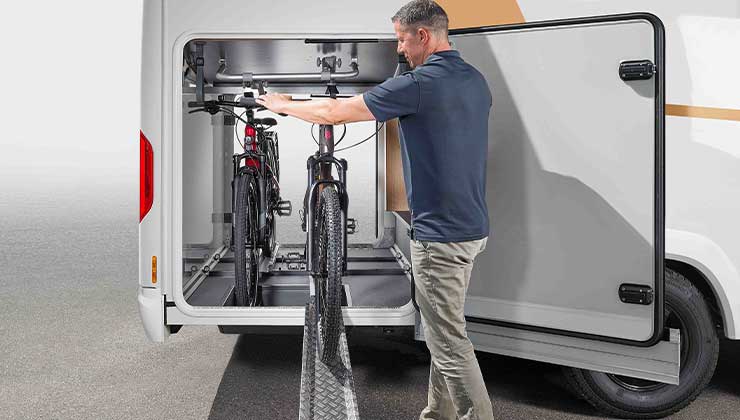 Ein Mann lädt zwei Fahrräder in den hinteren Stauraum eines Wohnmobils, wobei er den eingebauten, verschiebbaren Fahrradträger für einen einfachen Zugang und sicheren Transport nutzt.