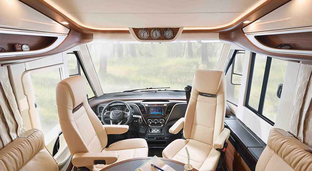Der Innenraum eines modernen Wohnmobils mit zwei hellbraunen Ledersesseln, einem kleinen runden Tisch und einer großen Frontscheibe, die eine verschwommene Szene im Freien mit Grünpflanzen zeigt. Das Armaturenbrett ist mit digitalen Bedienelementen und Holzakzenten versehen.