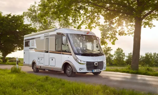 Ein weißes Wohnmobil fährt eine Landstraße entlang, die von grünem Gras und Bäumen umgeben ist, wobei das Sonnenlicht durch die Blätter fällt und der Himmel im Hintergrund klar ist.