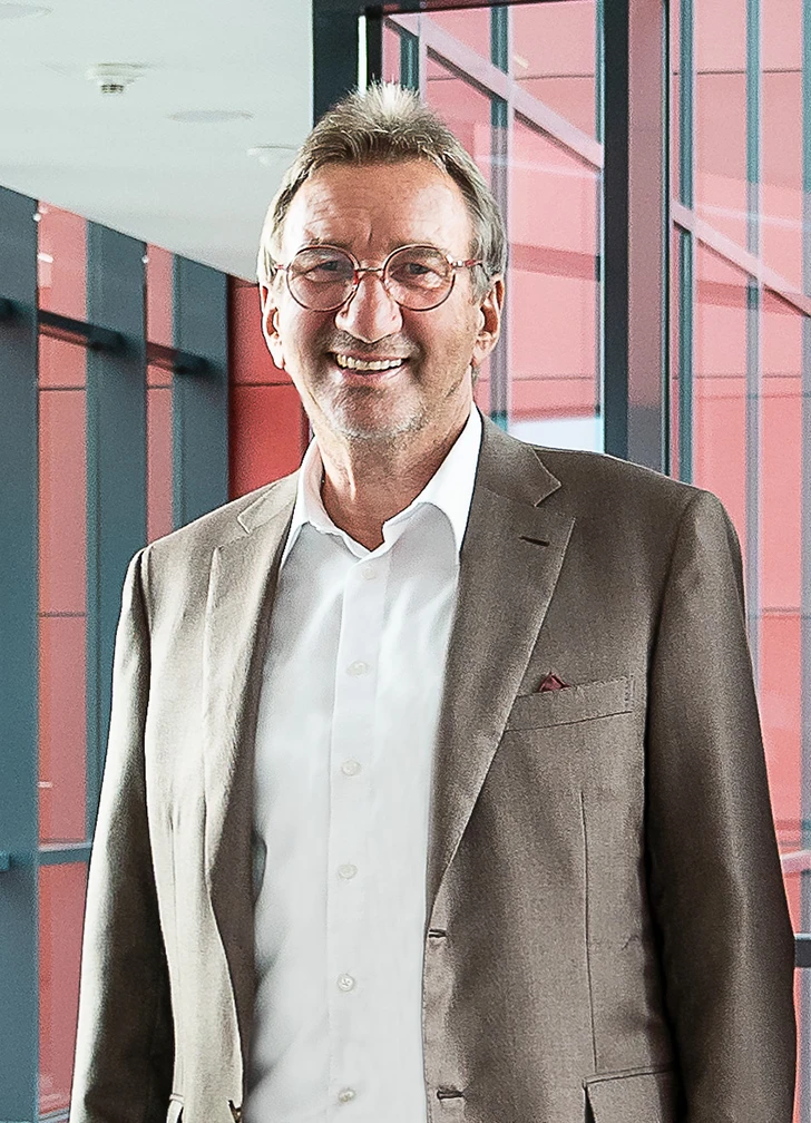Ein lächelnder Mann mit Brille, beigefarbenem Anzug und weißem Hemd steht in einem Innenraum vor großen Fenstern mit roter und schwarzer Verkleidung im Hintergrund.
