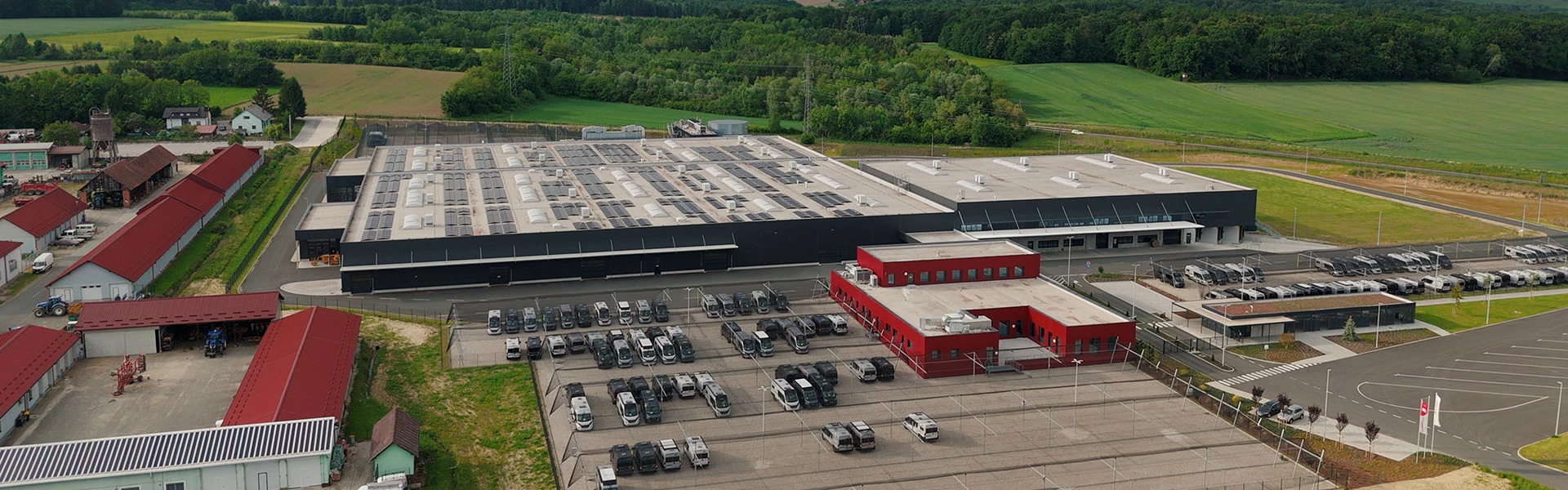 Luftaufnahme einer großen Industrieanlage mit einer grauen Lagerhalle, einem roten Bürogebäude und einem Parkplatz voller Autos, umgeben von grünen Feldern und Bäumen.